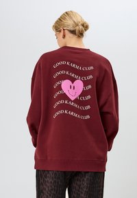 Bordeaux sweatshirt met een grote roze hartafbeelding met een glimlachend gezicht, omringd door herhaalde tekst met "GOOD KARMA CLUB" in het wit.