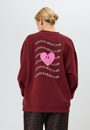 Bordeauxrotes Sweatshirt mit einem großen pinken Herzmotiv mit einem Smiley-Gesicht, umgeben von wiederholtem Text, der "GOOD KARMA CLUB" in Weiß sagt.