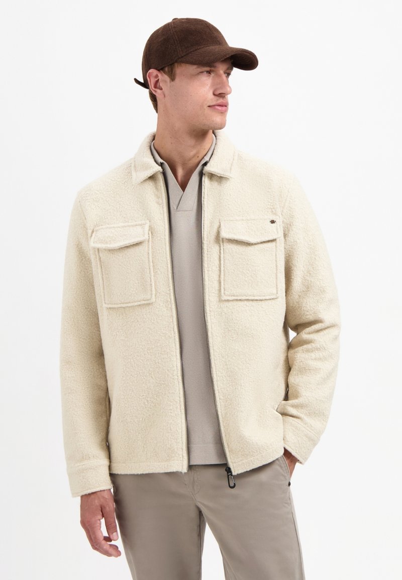 Fleece jacket in roomkleur met twee borstzakken, ritssluiting en een zachte textuur. Draagt over een beige top en gecombineerd met lichte broek. Bruine pet.