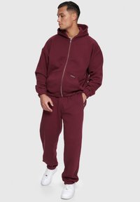 Mørkerød sæt bestående af sweatshirt og sweatpants, med en hættetrøje med lynlås, sidelommer og manchetter, parret med hvide sneakers.