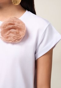 Camiseta blanca de manga corta con un gran adorno de flor de tela nude texturizada en el lado izquierdo del pecho.