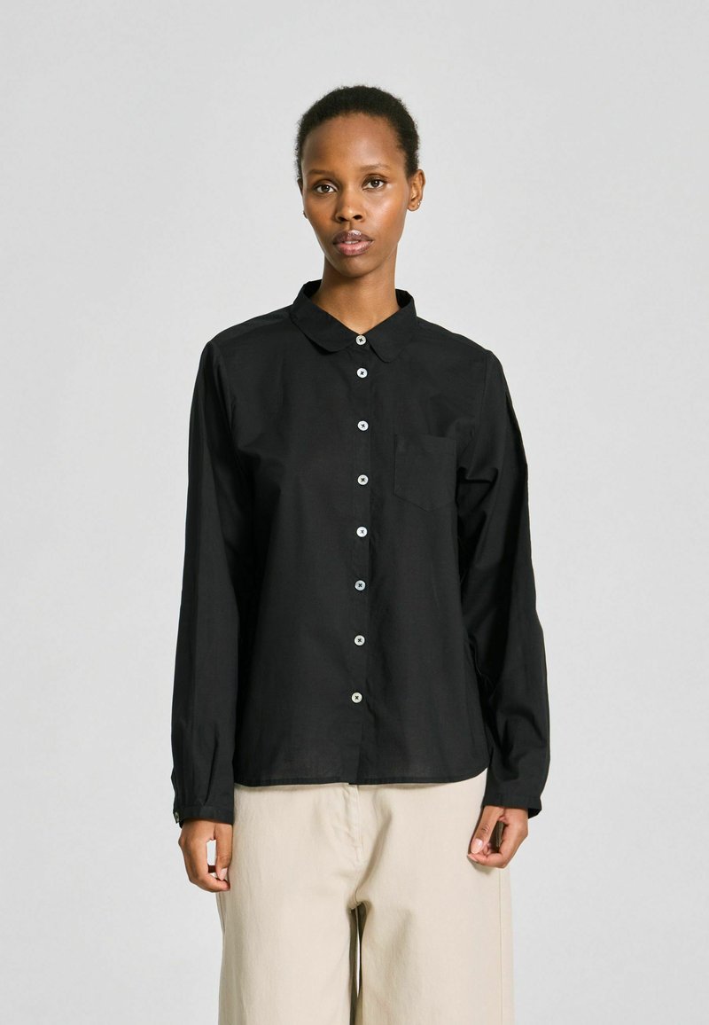 Personne debout, portant une chemise noire à manches longues avec une poche poitrine et un pantalon large beige clair, fond neutre.