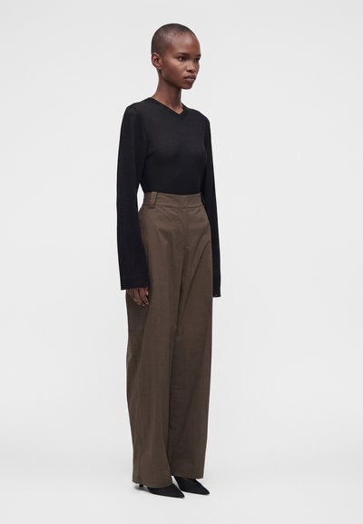 Femme aux cheveux courts portant un haut noir à manches longues, un pantalon marron taille haute à jambes larges et des chaussures noires pointues, debout de profil.