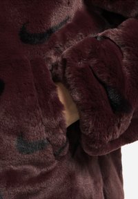 Burgundy faux fur-jacka med mjuk textur, med svarta Nike-loggor, en avslappnad passform och en mysig design med synliga handfickor.