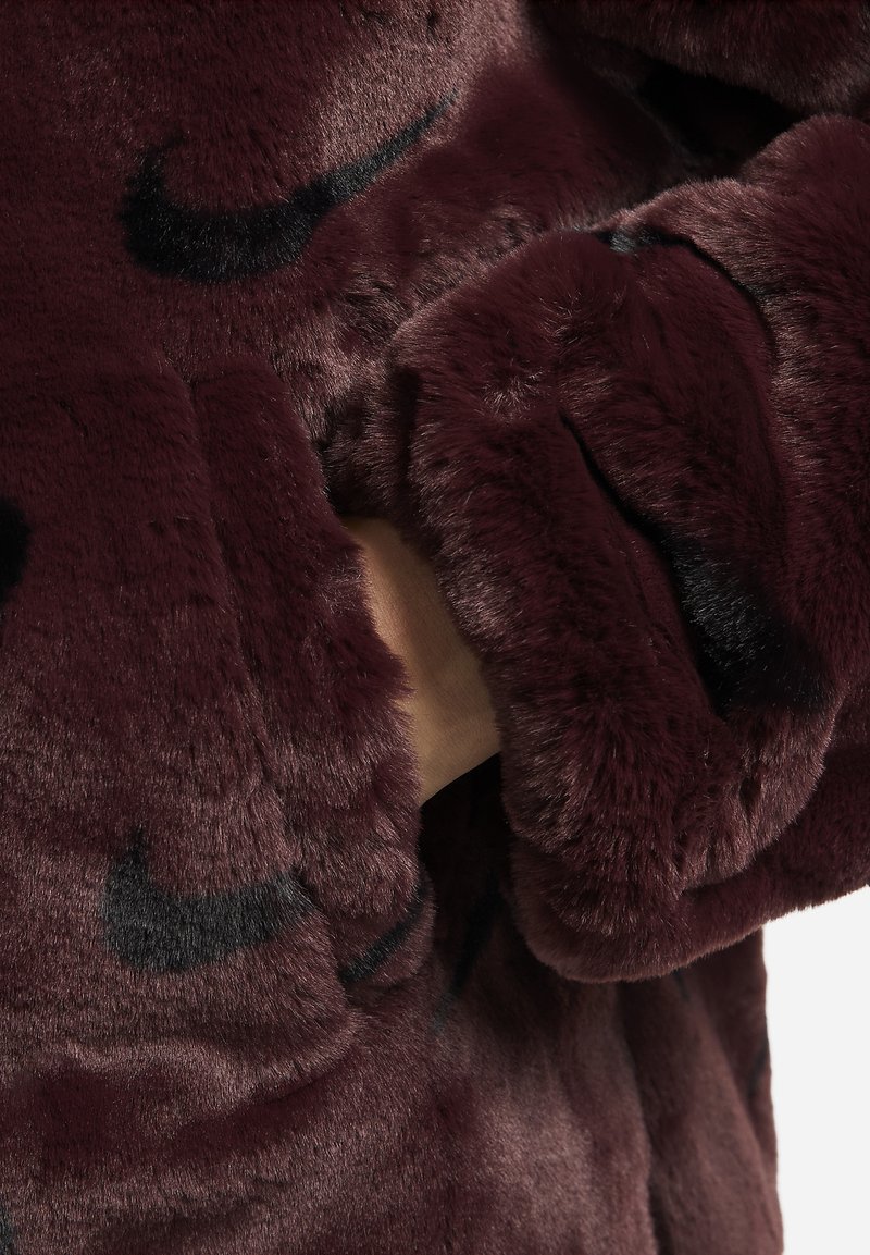 Veste en fausse fourrure bordeaux avec une texture douce, ornée de logos Nike noirs, à coupe décontractée et au design confortable avec des poches visibles.