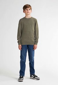 Zelený pletený svetr s kulatým výstřihem, doplněný modrými džínami a černo-bílými teniskami. Hladká textura, ležérní střih.