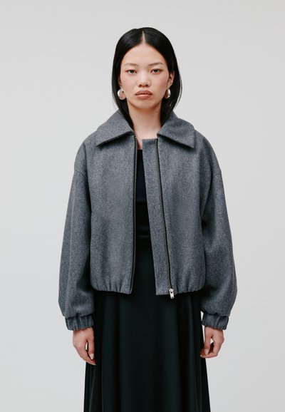 Graue, kurze Jacke aus strukturiertem Stoff mit breitem Kragen, Reißverschluss und gerafften Ärmeln, kombiniert mit einem schwarzen Kleid.