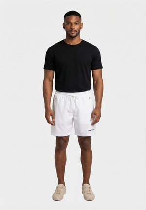 Hombre de pie vestido con camiseta negra, pantalones cortos blancos con cordón y bolsillos con cremallera, y zapatillas beige sobre fondo blanco liso.