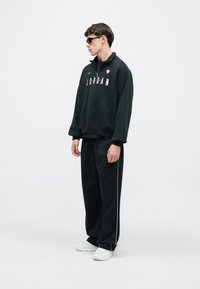 Μαύρο φούτερ με το κείμενο "JORDAN", λαιμόκοψη γύρω και φερμουάρ στο γιακά. Συνδυάζεται με μαύρο παντελόνι με ρίγες στα πλαϊνά και λευκά αθλητικά παπούτσια.