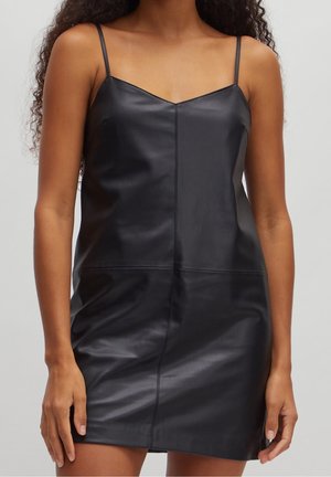 Kvinde iført en ærmeløs sort læder minikjole med tynde stropper, stående mod en ensartet lys grå baggrund.