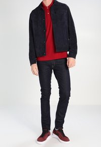 Granatowa zamszowa kurtka na czerwonej koszulce polo, zestawiona z ciemnymi dżinsami i bordowymi trampkami z białymi podeszwami. Strój ma casualowy charakter.