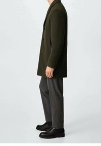 Donkergroene dubbelzijdige jas met een notch-kraag en twee knopen, gecombineerd met grijze broek en zwarte enkellaarsjes. Gladde stoftextuur.