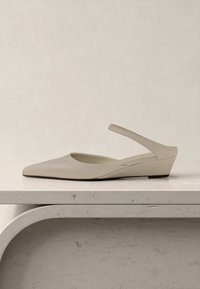 Chaussure beige à bout pointu avec bride arrière et petit talon compensé, présentée sur une surface en pierre claire texturée, sur un fond clair.