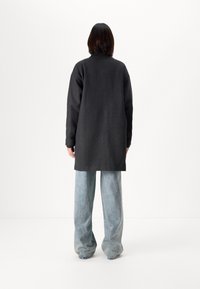 Lang svart grå frakk med høy krage, drop shoulders og rett silhuett, sammen med lyseblå vide jeans.