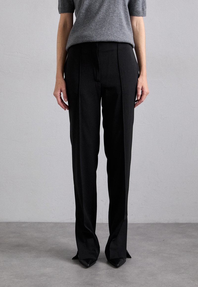 Black tailored trousers with a straight-leg design, featuring front pleats and a matte finish. Worn with a short-sleeve gray sweater.