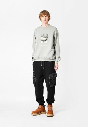 Sudadera gris con un diseño gráfico en el frente, combinada con pantalones cargo negros con cremalleras y bolsillos, y botas marrones.