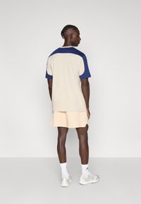 Bež košulja kratkih rukava s plavim naglascima na ramenima i okruglim izrezom, uparena sa svijetlo bež kratkim hlačama i bijelim tenisicama.