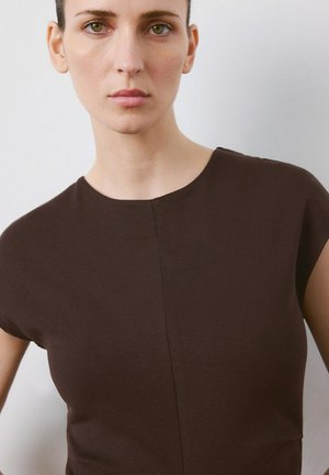 Giovane donna con pelle chiara e occhi verdi che indossa una maglia marrone scuro aderente a maniche corte su uno sfondo bianco uniforme.