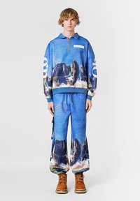 Sudadera azul con media cremallera y estampado de paisaje montañoso, combinada con pantalones azules a juego que presentan un diseño similar y perneras anchas. Botas marrones.