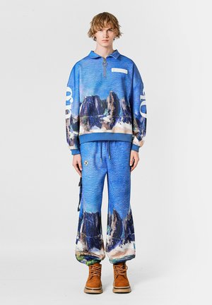 Sudadera azul con media cremallera y estampado de paisaje montañoso, combinada con pantalones azules a juego que presentan un diseño similar y perneras anchas. Botas marrones.