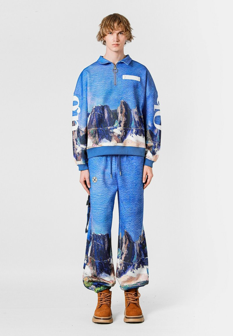 Sudadera azul con media cremallera y estampado de paisaje montañoso, combinada con pantalones azules a juego que presentan un diseño similar y perneras anchas. Botas marrones.