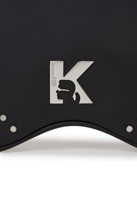Accesoriu din piele neagră cu un logo metalic argintiu în formă de „K”, având un contur. Textură netedă cu margini rotunjite și cuie metalice.