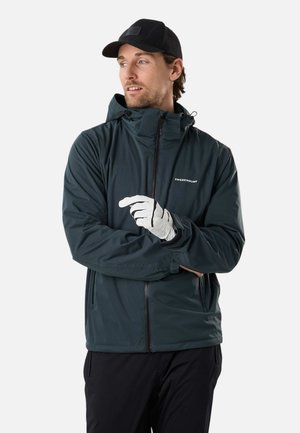 Mand iført sort kasket, mørkeblå Swedemount jakke med hætte, hvid handske og sorte bukser, kigger mod venstre med armene over kors.