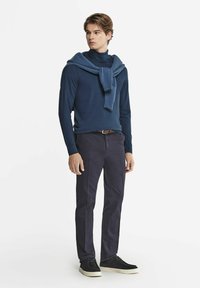 Marinblå långärmad skjorta med en matchande tröja draperad över axlarna, kombinerad med mörka byxor och svarta sneakers med vita sulor.