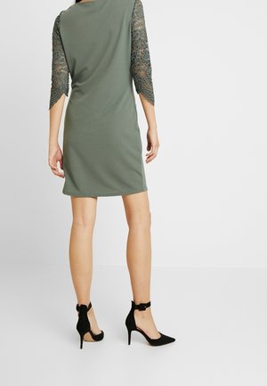 Frau steht mit dem Rücken zur Kamera, trägt ein knielanges olivgrünes Kleid mit dreiviertellangen Ärmeln aus Spitze und schwarze, hochhackige Schuhe mit Knöchelriemen.