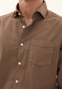 Brun button-up skjorta i lätt tyg, med spetsig krage, bröstficka och fem vita knappar fram. Slät textur.