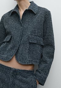 Giacca corta blu navy testurizzata con colletto, due tasche frontali e un sottile motivo a tessitura. Tessuto liscio e vestibilità sartoriale.