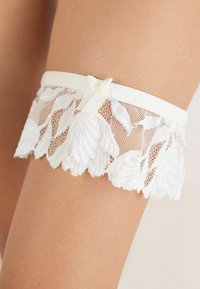 Vit spetsstrumpband med vågiga kanter och blommönster, med en satinrosett som detalj på bandet, designat för att passa bekvämt på låret.