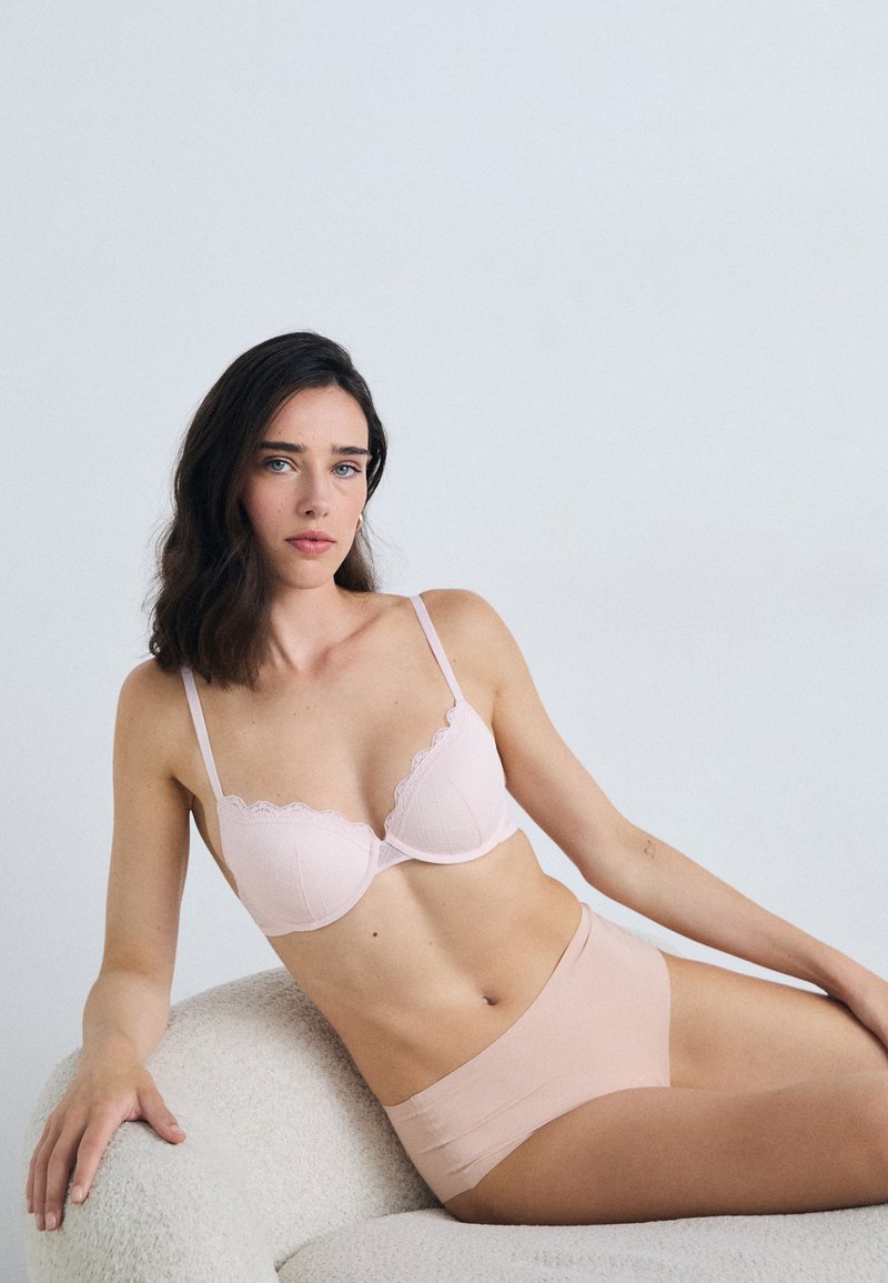 Lichtroze bh met golvende kantafwerking en gestructureerde cups, gecombineerd met naadloze beige slips, die de gladde stof en het minimalistische ontwerp benadrukken.