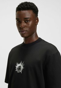 T-shirt en coton noir avec un col rond, des manches courtes et un graphique sur le devant représentant un visage souriant avec des accents en argent.