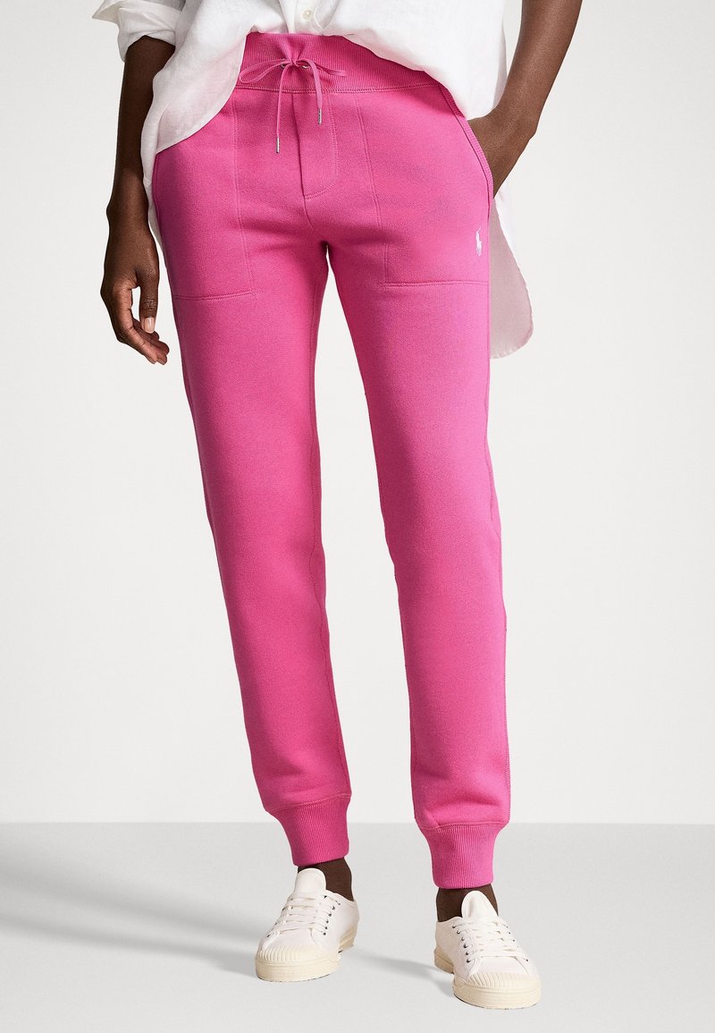 Rosa sweatpants med dragsko i midjan, två framfickor och ribbade muddar, parat med vita sneakers på en ljusgrå bakgrund.