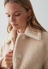 Giacca sherpa beige chiaro con una superficie texturizzata, grande colletto e due bottoni frontali. Tessuto morbido con un aspetto accogliente.