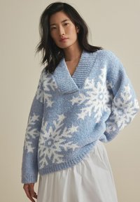 Maglione azzurro chiaro con motivi a fiocco di neve bianchi, texture lavorata a maglia, scollatura a V e orlo leggermente asimmetrico.