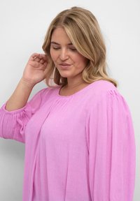 Lyserød, tekstureret bluse med lange ærmer og rund halsudskæring. Har bløde samlinger og små, hævede prikker i hele stoffet.