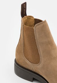 Tan suede ankle boot featuring textured elastic side panels, stitched accents, and a dark brown rubber sole. Branded pull tab included.