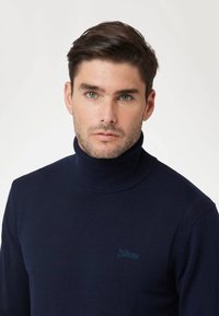 Maglione a collo alto in maglia navy con texture a coste, caratterizzato da un piccolo logo di colore blu chiaro. Design aderente con collo alto.
