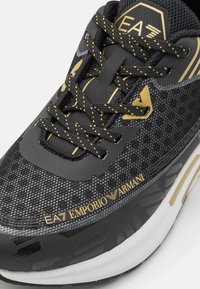 Schwarzer Sportschuh mit strukturiertem Obermaterial, goldenen Akzenten und kontrastierenden Schnürsenkeln. Verfügt über auffällige Markenkennzeichnung und eine gepolsterte Sohle.