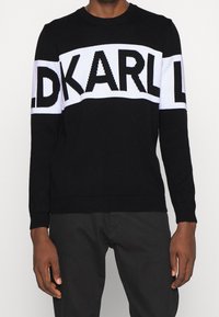 Mustavalkoinen neulottu neule, jossa on paksut "KARL"-kirjaimet rinnassa ja hihoissa, pyöreä pääntie ja resorireunaiset hihansuut.