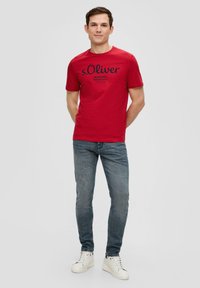 Rode katoenen t-shirt met zwart bedrukte logo en tekst, in combinatie met slim-fit blauwe jeans en witte sneakers.