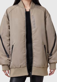 Beige Bomberjacke mit Reißverschluss, gerippten Bündchen und zwei seitlichen Reißverschlüssen. Verfügt über eine dezente Textur und einen kleinen Stoffpatch auf der Brust.