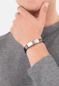 Pulsera de cuero negro con un cierre de metal plateado cepillado y dos rayas negras de acento. Lleva puesta en una mano sobre un suéter gris.