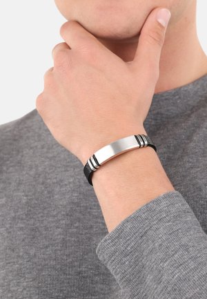 Pulsera de cuero negro con un cierre de metal plateado cepillado y dos rayas negras de acento. Lleva puesta en una mano sobre un suéter gris.