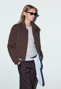 Giacca marrone trapuntata con colletto alto, camicia grigia sotto, e maglione azzurro chiaro annodato in vita, abbinati a pantaloni scuri e occhiali da sole neri.