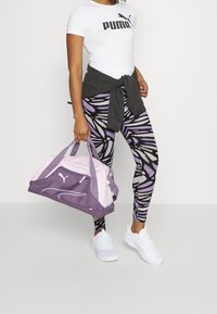 Witte Puma t-shirt, grijze sweater om de taille geknoopt, gemusterde paarse leggings en een paarse en roze sporttas. Witte sneakers.