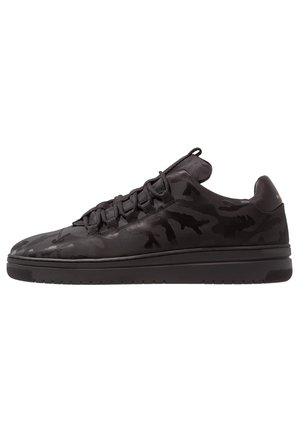 Zapatilla negra de caña baja con un sutil patrón de camuflaje, cordones negros y suela gruesa de goma sobre fondo blanco.