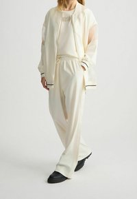 Kremová sportovní bunda s průhlednými rukávy a černými akcenty, spárovaná s volnými krémovými kalhotami a robustními černými botami.